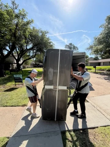 Two movers transporting upright piano with protective covering on residential driveway