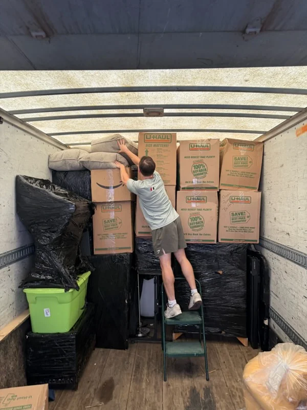 Moving truck interior with stacked cardboard boxes, wrapped furniture, and plastic-protected items secured for transport