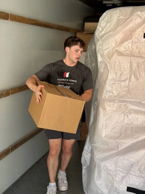 Moving crew member carrying cardboard box with furniture wrapped in protective plastic sheeting