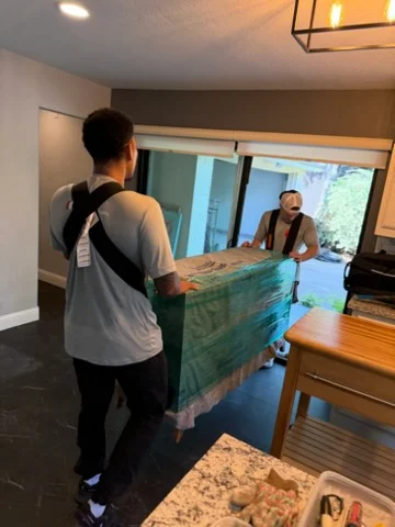 Movers carrying wrapped sofa through residential interior with wood flooring