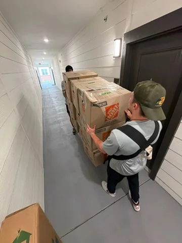 Movers carrying stacked cardboard boxes through narrow hallway with white walls